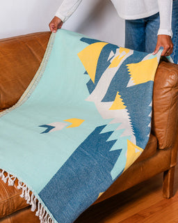 A person holds up a woven throw blanket with a Southwestern mountain pattern, draped over a brown leather couch in a living room setting.