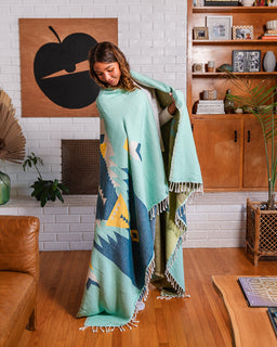 A woman stands in a living room wrapped in a large woven throw blanket. The blanket is mint green with a Southwestern-style geometric pattern in blue, yellow, and beige, and has white fringe on the edges.