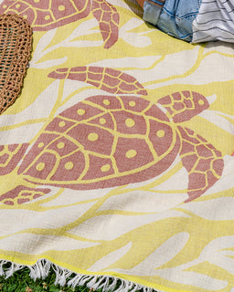 A close-up of a woven blanket or towel with a yellow and white background and a large pink sea turtle print, laid outdoors on grass with part of a woven mat and blue jeans visible.