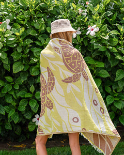 A woman seen from behind wearing a beige crochet bucket hat and wrapped in a yellow and brown sea turtle patterned blanket, standing in front of a green leafy garden wall with white flowers.