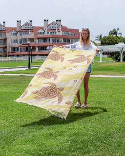A woman standing on a grassy lawn holds up a large beach towel  with a yellow background and brown sea turtle pattern. Apartment buildings are visible in the background.