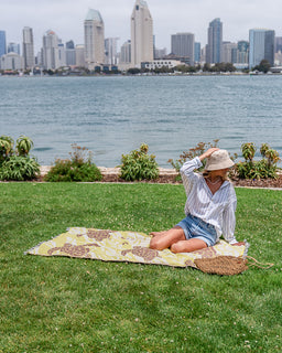 A woman sits on a yellow and brown patterned picnic blanket on a green lawn, with a waterfront city skyline in the background. She is wearing a white striped shirt, denim shorts, and a beige sun hat.
