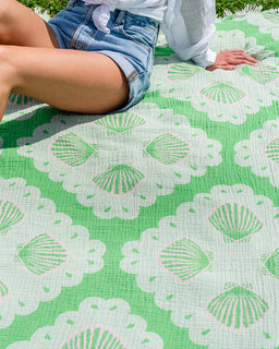 A close-up of a green and white patterned picnic blanket with a scalloped shell design, with a person sitting on it wearing denim shorts and a white shirt.