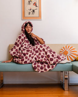 A woman reclining on a light blue bench wrapped in a plush hooded blanket with a burgundy floral pattern, in a modern room with a wooden floor and framed wall art.
