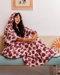 A woman sits on a beige couch wrapped in a plush hooded blanket with a pink and burgundy floral pattern, in a brightly lit living room.
