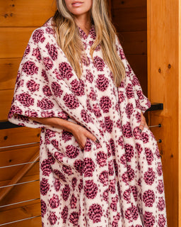 A woman wearing a cream-colored robe with a dark red floral pattern, standing in front of a wooden wall.