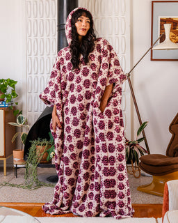 A woman wearing a plush hooded kaftan robe with a dark purple and cream floral pattern, standing in a modern living room setting.