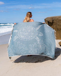 Person holding a beach towel with turtle design on a sandy beach.