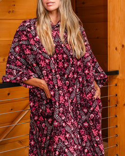 A woman wearing a loose wearable blanket with a dark purple and pink floral pattern, standing in front of a wooden wall.