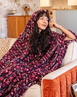 A woman with long dark hair sits on a cream couch wrapped in a plush hooded blanket featuring a pink, purple, and black floral and abstract pattern. The setting is a cozy living room with warm lighting.