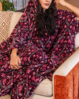 A woman wrapped in a plush hooded blanket with a dense floral and leaf pattern in deep magenta, burgundy, and gray, sitting on a cream-colored couch with a rust-orange armrest.