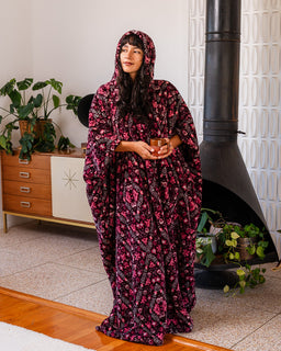 A woman wearing a hooded, floor-length robe with a pink and black floral pattern, holding a small brown cup in a home interior with a fireplace and plants.
