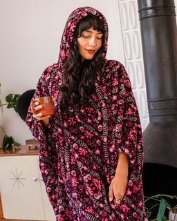 A woman wearing a plush hooded blanket or poncho with a pink, red, and black floral pattern, holding a small orange mug in a cozy indoor setting.