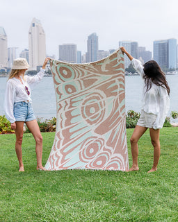 Two women standing on a grassy lawn hold up a large woven towel with a bohemian abstract pattern in sage green, dusty pink, and cream, with a city skyline and water in the background.