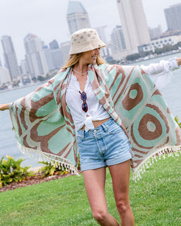 A woman stands outdoors on a grassy area near water with a city skyline in the background, wearing a large towel with mint green, brown, and cream abstract pattern over a white top and denim shorts.