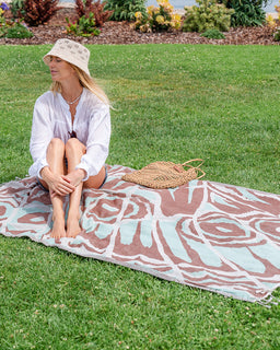 A woman sits barefoot on a large patterned beach towel spread out on a green lawn, wearing a white long-sleeve shirt and a beige crocheted sun hat. A woven straw bag rests beside her on the towel.