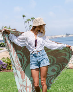A woman outdoors by the water holds up a large patterned towel with both hands. She is wearing a white tied blouse, denim shorts, and a woven hat.