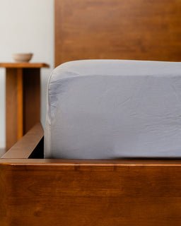 Wooden bed frame with a gray fitted sheet in a minimalistic room.