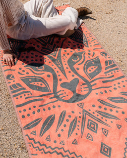 Person sitting on a orange patterned towel with black designs on a sandy surface
