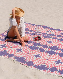 Person sitting on a colorful beach towel with a straw hat, white shirt, and shorts.