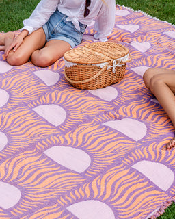 A purple and orange patterned picnic blanket spread on grass, with a wicker picnic basket in the center and parts of two people visible sitting and reclining on the blanket.