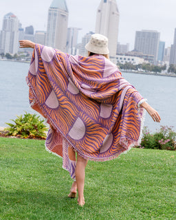 A woman stands on a grassy lawn with a city skyline and water in the background, wearing a large patterned wrap or shawl draped over her body. The fabric has an orange, purple, and pink abstract design with fringe trim.