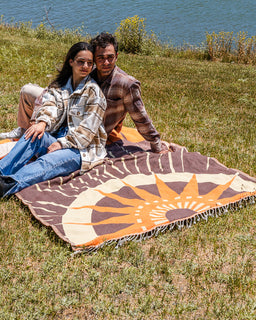 A couple sits on a large woven blanket with a sunburst pattern, placed on grass near a body of water. The blanket is brown, cream, and orange, and the people are wearing casual outdoor clothing.