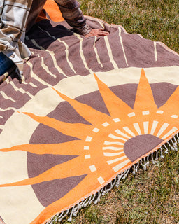 A large woven blanket with a sunburst design is laid out on grass. The blanket features a cream background with a prominent orange and dark brown sun motif, and a striped border.