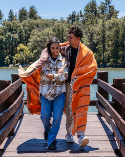 A couple walking arm in arm on a wooden dock by the water, wrapped together in a large orange blanket with black stripes. The woman wears a beige and white plaid flannel shirt with blue jeans, and the man wears a black t-shirt and light-colored pants.