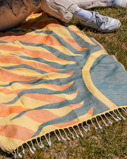 A colorful outdoor blanket with a wavy abstract pattern in orange, yellow, and teal, laid on grass with a person's legs and sneakers visible at the top edge.