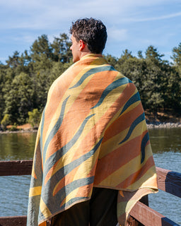 A man standing on a wooden dock by a lake, wrapped in a large striped blanket or throw with wavy bands of orange, yellow, blue, and beige.