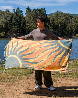 A man standing outdoors by a lake holds up a large woven blanket or towel featuring a stylized sun design in orange, yellow, and teal, with fringe on one edge.