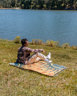 A man sits on a colorful patterned blanket on a grassy hill overlooking a large lake, wearing a brown plaid shirt and beige pants.