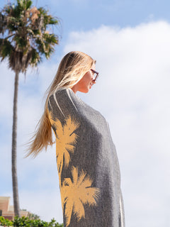 Woman wearing a gray poncho with palm tree patterns, standing outdoors with palm trees and blue sky in the background.