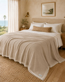 A beige textured throw blanket with fringe trim draped over a bed in a warm, sunlit bedroom. The bed is styled with white linens, a tan pillow, and a window with sheer curtains reveals a view of the ocean.