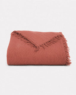 A folded textured throw blanket in a muted terracotta or dusty rose color, featuring fringed edges, set against a plain white background.
