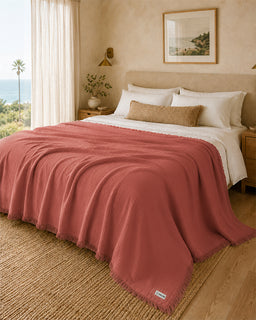 A textured dusty rose throw blanket with fringe is draped over a bed in a warm, modern bedroom setting with beige walls, wooden floors, and a window overlooking the ocean.