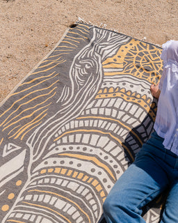 Person sitting on a patterned towel with geometric design on a sandy surface