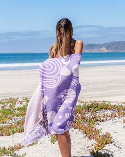 A woman standing on a sandy beach wrapped in a lavender and white patterned towel or wrap, with the ocean and blue sky in the background.