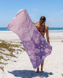 A woman on a sandy beach holds up a large lavender and purple patterned beach towel with abstract wave and shell-like motifs, with the ocean and blue sky in the background.