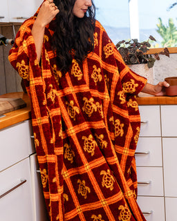 A woman wrapped in a plush blanket standing in a kitchen. The blanket is dark red-burgundy with a mustard yellow grid pattern and sea turtle motifs.