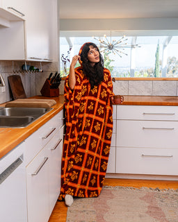 A woman wearing a long, loose-fitting wearable blanket with an orange and black geometric pattern stands in a modern kitchen, holding a small brown mug.