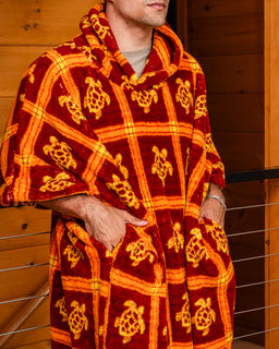 A man wearing a plush hooded blanket wrap in a warm wood-paneled setting. The blanket features a bold red and orange plaid pattern with yellow sea turtle motifs.