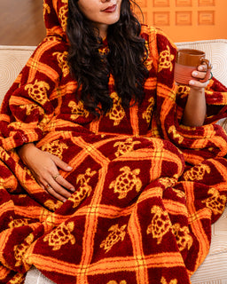 A woman reclining on a couch wrapped in a plush hooded blanket. The blanket is deep red with a mustard yellow grid pattern and turtle motifs.