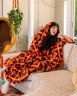A woman sits on a patterned sofa wrapped in a plush throw blanket featuring a bold red and orange geometric design with skull motifs. The setting is a cozy, modern living room with a hanging plant and warm natural light.
