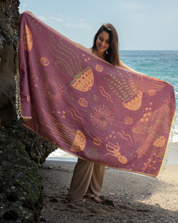 Women holding a maroon beach towel with a marine life design, including jellyfish and sea anemones, with twisted tassels at the bottom at the beach