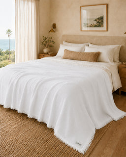 A white textured throw blanket with fringe trim draped over a bed in a warm, neutral-toned bedroom. The bed is styled with white and beige pillows, set against a beige headboard and wall with a window showing a view of the ocean and palm trees.