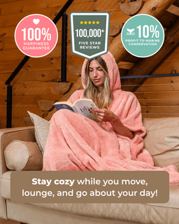 A woman sits on a beige sofa wrapped in a soft pink hooded blanket, reading a book in a rustic wood-paneled room. Promotional text and badge graphics appear around the scene, including 'Stay cozy while you move, lounge, and go about your day!' and trust badges about reviews and happiness guarantee.