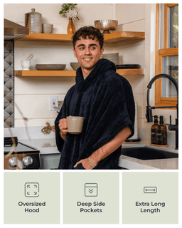 A man wearing a plush oversized bathrobe stands in a kitchen holding a mug. The robe is dark navy blue and has a hood.