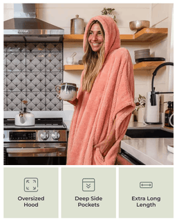 A woman in a kitchen wearing a oversized terracotta pink hooded towel robe with side pockets, holding a mug. Three feature callout panels at the bottom highlight the oversized hood, deep side pockets, and extra long length.